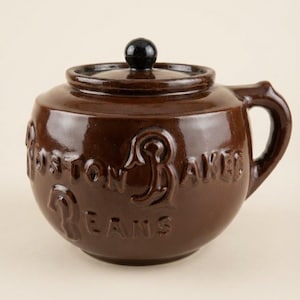 May include: A round, dark brown glazed ceramic bean pot with a matching lid featuring a black knob. The exterior is embossed with the text "BOSTON BAKED BEANS" in raised lettering. The pot includes a single side handle and a wide, circular base.
