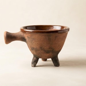 May include: A brown terracotta bowl with a smooth, glazed rim and a matte, textured exterior. The bowl features a single horizontal handle on one side and is supported by three short, sturdy legs, giving it a distinct tripod base structure.
