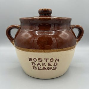 May include: A two-toned ceramic bean pot with a glossy brown upper section and a matte cream lower section. It features two side handles, a matching lid with a knob, and the text BOSTON BAKED BEANS embossed in brown on the front of the pot.