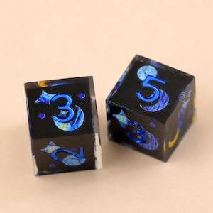 May include: Two six-sided dice featuring a dark, swirling blue and yellow pattern reminiscent of a starry night sky. The numbers three, two, and five are engraved in a metallic blue finish on the faces, accented by small star and moon shapes.