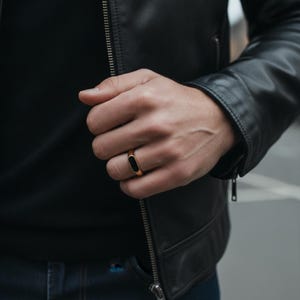 May include: A gold-toned ring featuring a smooth, elongated black oval inlay on the front surface. The band has a polished finish and is worn on a finger, positioned against a black leather jacket and a dark shirt, showcasing a sleek design.