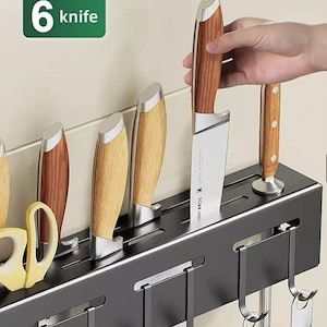 Puede incluir: Un organizador de cocina de pared negro con ranuras para cuchillos de mango de madera y virolas de metal plateado, junto con varios ganchos integrados para colgar utensilios. Una etiqueta rectangular verde muestra el texto 6 knife.