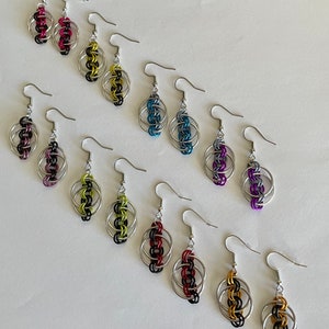 Pode incluir: Uma coleção de brincos artesanais com um design de malha de corrente. Cada brinco apresenta uma armação de metal prateado com detalhes coloridos de malha de corrente em várias cores, incluindo rosa, preto, amarelo, azul, roxo, vermelho e verde. Os brincos têm um fecho de gancho.