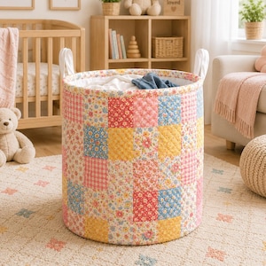 May include: A cylindrical laundry hamper featuring a quilted patchwork design with various floral and checkered patterns in shades of pink, yellow, and blue. The hamper includes two white fabric handles attached to the top rim for easy carrying.