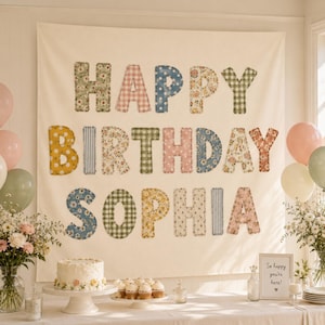 Op de afbeelding: Een grote wandhanger van gebroken witte stof met de tekst HAPPY BIRTHDAY SOPHIA in kleurrijke, bedrukte stoffen letters. Elke letter heeft een uniek patroon, waaronder bloemen, ruitjes, stippen en strepen in zachte kleurtinten.