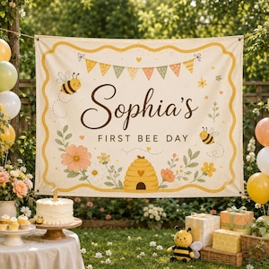 Könnte beinhalten: Ein rechteckiges Stoffbanner mit cremefarbenem Hintergrund und gelbem Wellenrand. Der Text lautet Sophia's FIRST BEE DAY in dunkelbrauner Schrift. Die Illustrationen zeigen zwei Bienen, einen Bienenstock und Blumen.