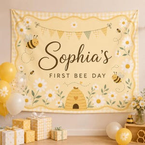 Könnte beinhalten: Ein rechteckiger Stoffhintergrund mit gelb-weißem Vichy-Karo-Rand, verziert mit Gänseblümchen, Bienen und einer Wimpelkette. In der Mitte steht der Text "Sophia's FIRST BEE DAY" in dunkelbrauner Schrift über einem Bienenstock.