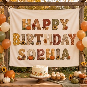 May include: A cream-colored fabric banner featuring the text HAPPY BIRTHDAY SOPHIA in large, patterned letters decorated with autumn motifs like pumpkins, sunflowers, and leaves. The banner is surrounded by orange and white balloons.