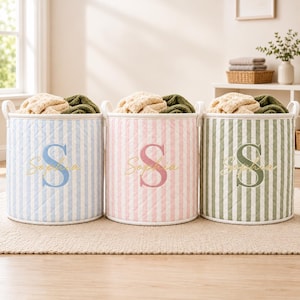 May include: Three cylindrical fabric storage baskets with quilted vertical stripes in light blue, soft pink, and sage green. Each basket features two side handles and is personalized with a large embroidered letter S and the name Sophia in script.