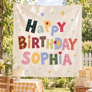 May include: A cream-colored fabric banner featuring the text "Happy Birthday Sophia" in large, multi-colored, patterned letters. The design includes various plaid, floral, and polka dot prints, accented by small stars, dots, and a red heart.
