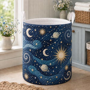 May include: A cylindrical laundry hamper featuring a quilted dark blue fabric exterior with a celestial pattern of gold and light blue suns, moons, planets, and stars. The hamper includes two white fabric handles and a white interior lining.