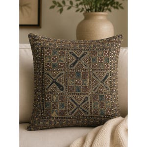 May include: A square throw pillow featuring a woven tapestry-style fabric with a repeating geometric pattern. The design consists of alternating dark brown, black, and muted blue squares and X-shaped motifs, accented with small white dotted details.