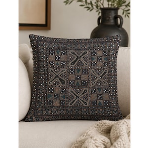 May include: A square throw pillow featuring a dark charcoal fabric base with an intricate, multi-colored embroidered pattern. The design consists of repeating geometric squares and cross motifs in shades of brown, blue, and white thread.