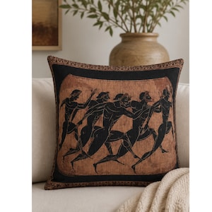 Pode incluir: Uma almofada quadrada com um design em preto e cor terracota inspirado na cerâmica grega antiga. O gráfico central mostra um grupo de cinco figuras em movimento, representadas em silhuetas pretas sobre um fundo texturizado de cor terracota.