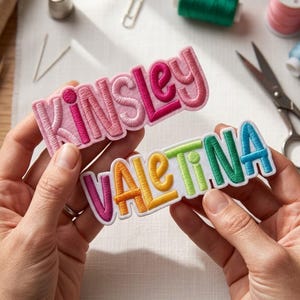 May include: Two embroidered name patches with white felt backing. The top patch features the name KINSLEY in shades of pink and magenta. The bottom patch features the name VALENTINA in a rainbow gradient of purple, orange, yellow, green, and blue.