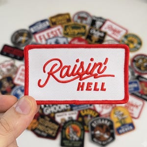 May include: A rectangular embroidered patch with a white background and a thick red border. The text "Raisin'" is stitched in red cursive script above the word "HELL" in red block capital letters, centered on the patch.