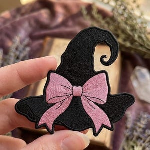 May include: An embroidered patch in the shape of a black witch hat with a curved top and a wide brim. The hat is decorated with a large, light pink bow centered at the base of the crown, featuring detailed stitching throughout the fabric design.