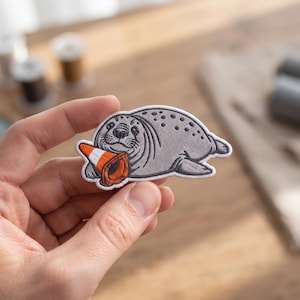 May include: An embroidered patch featuring a grey seal with dark spots, lying down and holding an orange and white traffic cone in its flippers. The patch has a white border and detailed black stitching to define the seal's facial features.