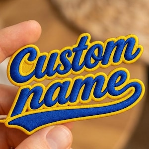 May include: An embroidered patch featuring the text Custom name in a bold, blue cursive script font. The lettering is outlined with a thick, bright yellow border, creating a high-contrast design with a textured, stitched fabric appearance.