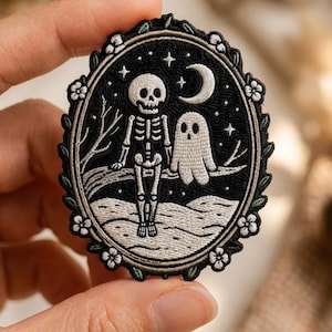 May include: An oval embroidered patch featuring a black background with a white skeleton and a white ghost sitting together on a tree branch. The scene includes a crescent moon, stars, and a floral border with small white flowers and green leaves.