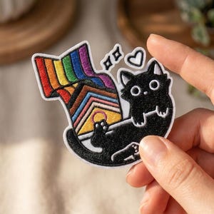 May include: An embroidered patch featuring a black cat with white eyes lying down next to a house-shaped structure. The house displays the Progress Pride flag colors, and a rainbow flag extends from the roof. Two small white hearts float above the cat.