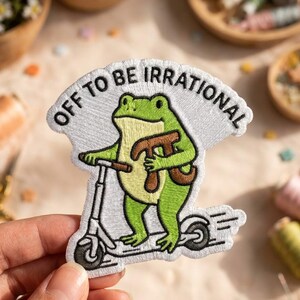 May include: An embroidered patch featuring a green frog riding a kick scooter and holding a brown pi symbol. The patch has a white background and includes the black embroidered text "OFF TO BE IRRATIONAL" arched across the top of the design.