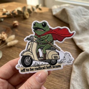 May include: An embroidered patch featuring a green frog wearing a red cape and brown boots, riding a cream-colored scooter. The frog carries a sword at its waist. Below the scooter, the text reads, "off to be radical left scum" in black letters.