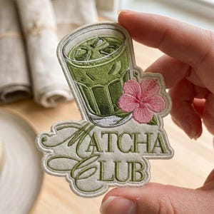 May include: An embroidered patch featuring a glass of iced green matcha tea with a pink hibiscus flower. The text MATCHA CLUB is stitched in olive green thread below the glass. The patch has a cream-colored background and a stitched border.