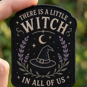 May include: A black embroidered patch featuring the text "THERE IS A LITTLE WITCH IN ALL OF US" in beige thread. The design includes a central witch hat, a crescent moon, small stars, and two sprigs of purple lavender leaves on the sides.