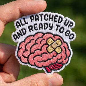 May include: An embroidered patch featuring a pink brain with a tan adhesive bandage on the surface. The text "ALL PATCHED UP AND READY TO GO" is stitched in black thread on a white background above the brain illustration with a white border.