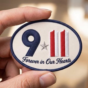 May include: An oval-shaped embroidered patch with a cream-colored background and a dark blue border. The design features the text 9-11 in dark blue and red, a small silver star, and the text Forever in Our Hearts in dark blue script lettering.