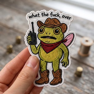May include: An embroidered patch featuring a green frog wearing a brown cowboy hat, brown cowboy boots, and a red bandana. The frog holds a black walkie-talkie and has small pink wings. The text "what the fuck, over" is embroidered in black above the frog.
