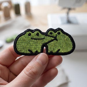 May include: An embroidered patch featuring two light green frogs with dark green outlines. The frogs are positioned side-by-side, with the left frog appearing to lean toward the right frog. The patch has a thick black border and a textured surface.