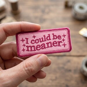 May include: A rectangular embroidered patch with a light pink background and a dark pink border. The text "I could be meaner" is stitched in dark pink thread, accompanied by four small star-like symbols placed around the lettering.