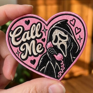 May include: A heart-shaped embroidered patch with a pink background featuring a black-hooded figure holding a pink telephone receiver. The text "Call Me" is stitched in cream-colored script. Small black hearts and stars decorate the pink field.
