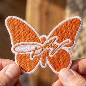 May include: An embroidered butterfly-shaped patch featuring a vibrant orange base with a white border. The word Dolly is stitched across the center in a white cursive script font, creating a distinct contrast against the orange fabric background.