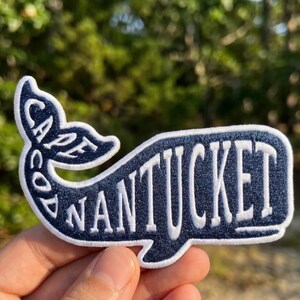 May include: A navy blue embroidered patch shaped like a whale with a white border. The word NANTUCKET is stitched in white across the body of the whale, and the words CAPE COD are stitched in white along the curved tail section of the patch.