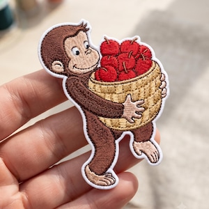 May include: An embroidered patch featuring a brown monkey character holding a tan woven basket filled with bright red apples. The design has a white border and detailed stitching throughout, depicting the monkey with a smiling expression.