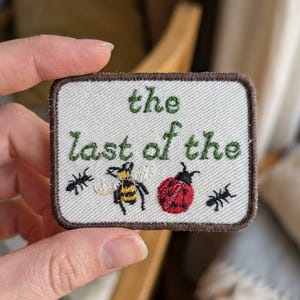 May include: A rectangular embroidered patch with a cream-colored background and a dark brown border. The text "the last of the" is stitched in green thread above a small embroidered bee, a red ladybug, and two black ants on the fabric surface.
