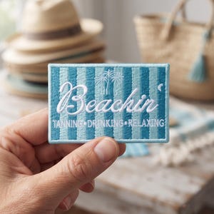 May include: A rectangular embroidered patch featuring vertical stripes in shades of light and dark blue. The text Beachin' is stitched in white script above the words TANNING • DRINKING • RELAXING, with a small white palm tree icon centered above.