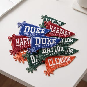 May include: A collection of triangular felt pennant patches in various colors including blue, red, green, and orange. Each patch features embroidered white university names such as Duke, Harvard, Baylor, and Clemson, with a decorative bow detail.
