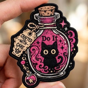 May include: An embroidered patch featuring a black cat inside a pink potion bottle. The bottle has a cork stopper, a tag reading "CRAFTED WITH CARE", and the text "Do It" above the cat. A small charm with a star hangs from the bottle neck.