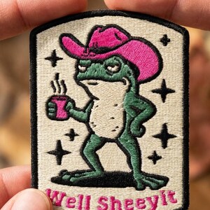 May include: A rectangular embroidered patch with a cream background and black border. It features a green frog wearing a bright pink cowboy hat, holding a pink mug with steam rising. Black star icons surround the frog, and the text Well Sheeyit is embroidered in pink at the bottom.