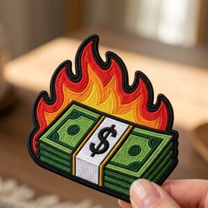 May include: An embroidered patch featuring a stack of green paper money bills wrapped in a white band with a black dollar sign symbol. The stack of money is engulfed in stylized flames colored in vibrant shades of red, orange, and yellow.
