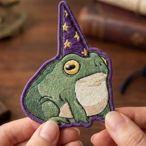 May include: An embroidered patch featuring a green frog with a light-colored belly, wearing a pointed purple wizard hat decorated with small yellow embroidered stars. The patch has a thick purple stitched border outlining the entire shape.