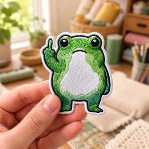 May include: An embroidered patch featuring a green frog with a white belly, large black eyes, and a dark outline. The frog is depicted with one arm raised, extending its middle finger. The patch has a white border and a textured, stitched surface.