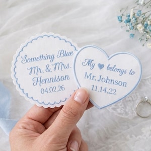 May include: Two white fabric patches with light blue embroidery. One circular patch with scalloped edges reads Something Blue Mr. & Mrs. Hennison 04.02.26. One heart-shaped patch reads My heart belongs to Mr. Johnson 11.14.22 with a blue heart icon.