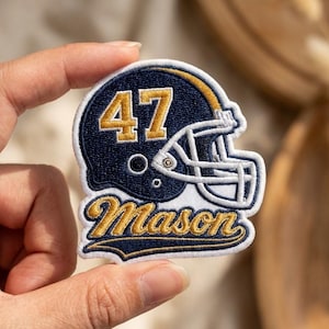 May include: An embroidered patch featuring a navy blue football helmet with a white face mask and the number 47 in gold. Below the helmet, the name Mason is stitched in gold cursive script with a navy blue outline on a white background.