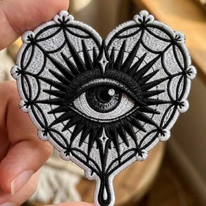 May include: A heart-shaped embroidered patch featuring a central black and white eye surrounded by radiating black spikes. The background of the heart is filled with a white web-like pattern, and the bottom of the heart has a single black drip.
