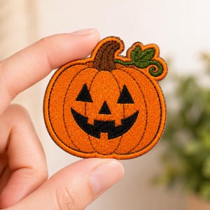 May include: An embroidered patch shaped like a jack-o'-lantern pumpkin. The pumpkin is bright orange with a brown stem, a green leaf, and a black stitched face featuring triangular eyes, a triangular nose, and a jagged, wide, toothless grin.